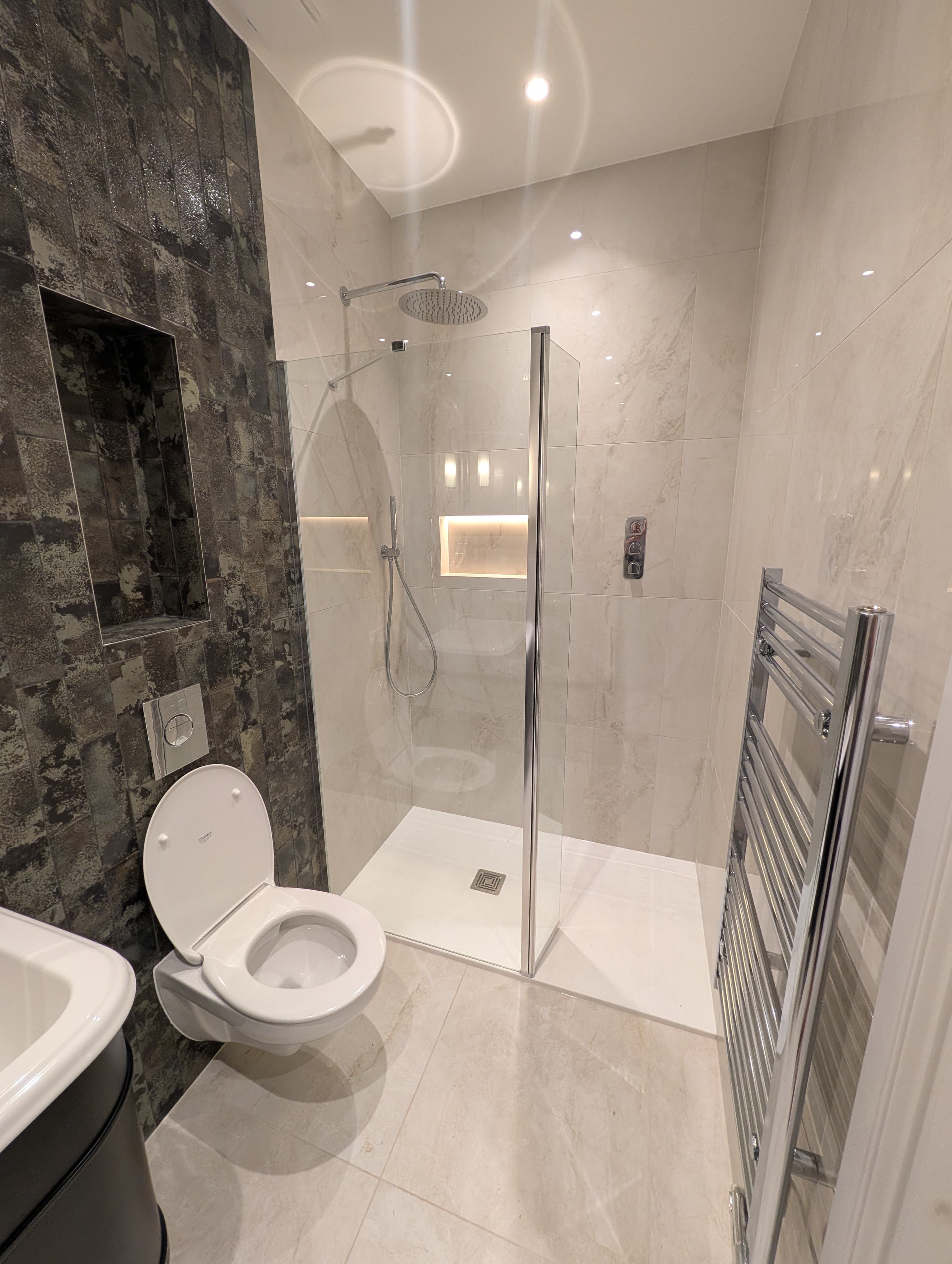 Bath raised on a tiled step below a textured tile wall, glass shower screen with chrome riser rail alongside