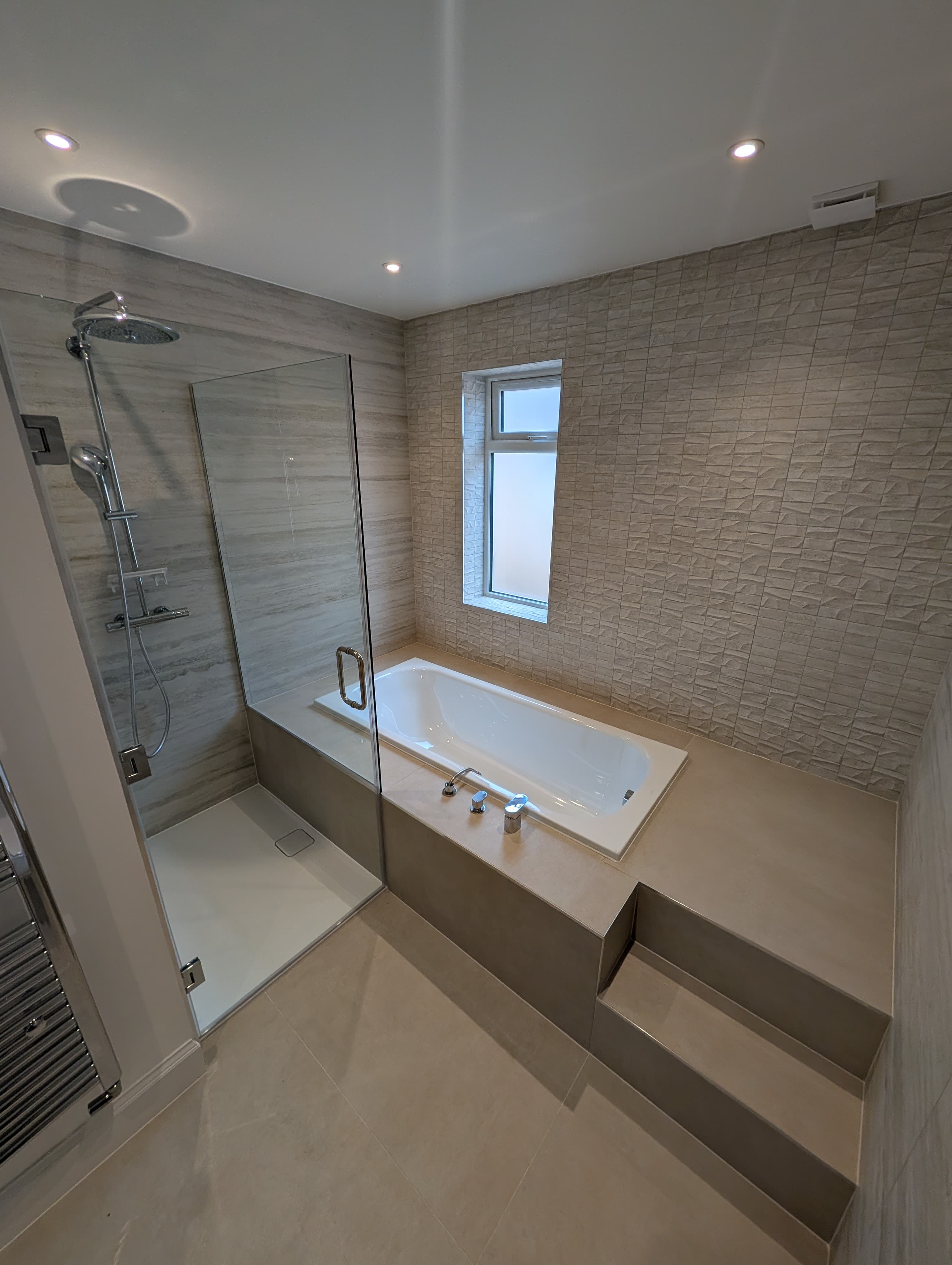 Corner shower with black recessed niche and ceiling-mounted rainfall head beside a bath with floor-standing chrome filler tap, below a window in a textured tile bathroom