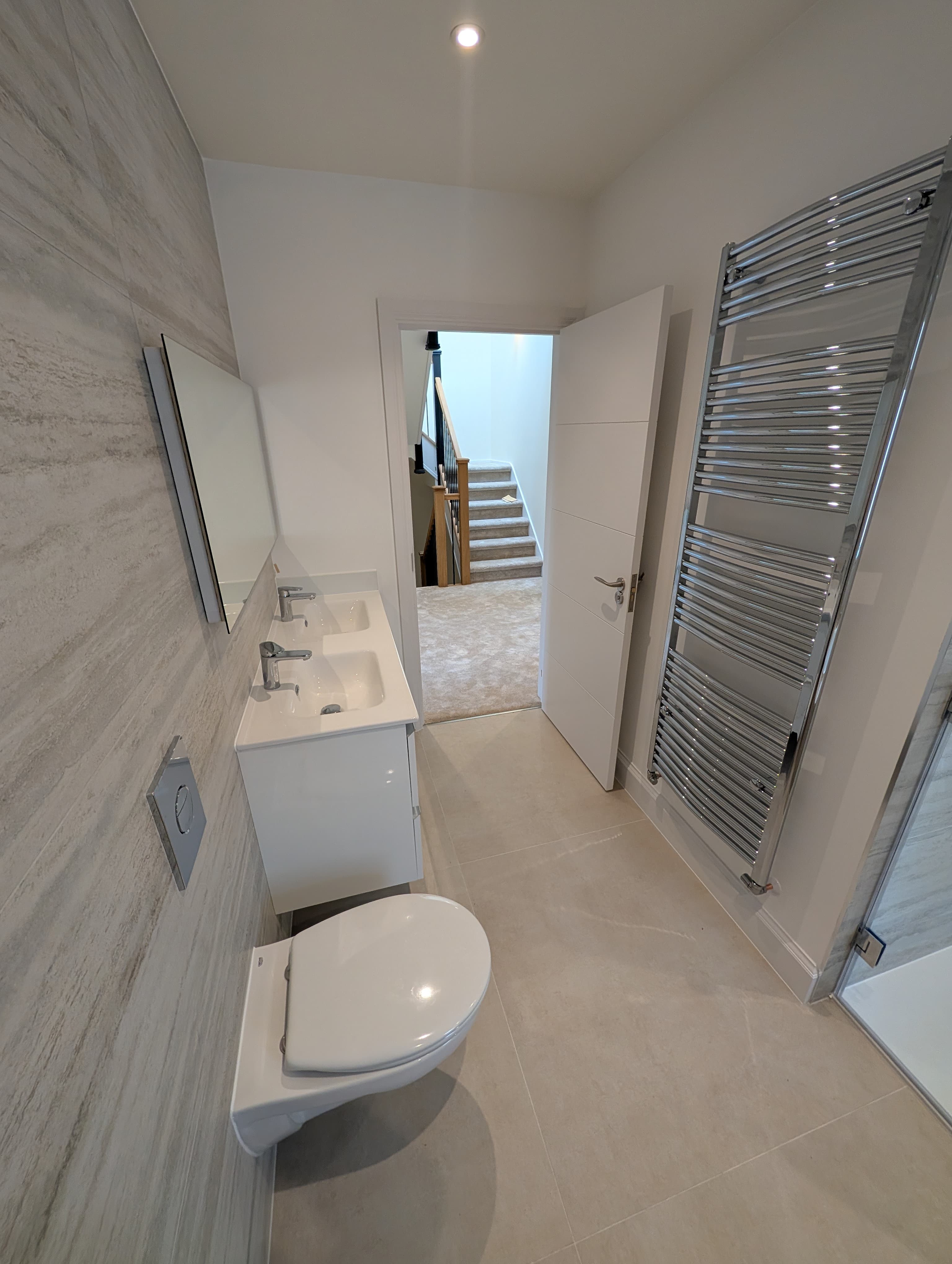 Wall-hung toilet and vanity mirror beside a chrome ladder heated towel rail, doorway opening onto a staircase landing