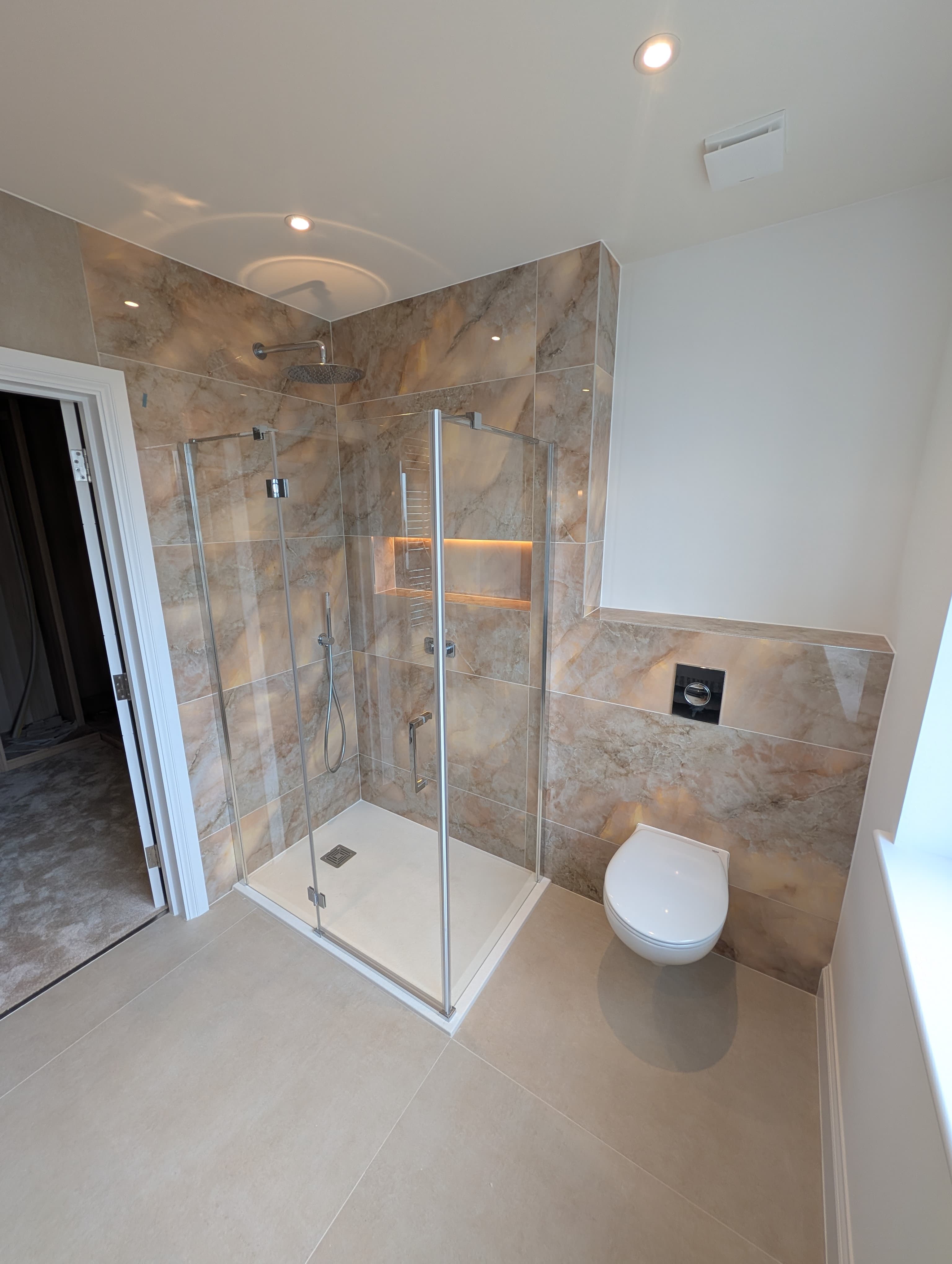Corner glass shower with warm marble-effect tiling, backlit recessed niche, chrome fittings and a wall-hung toilet alongside