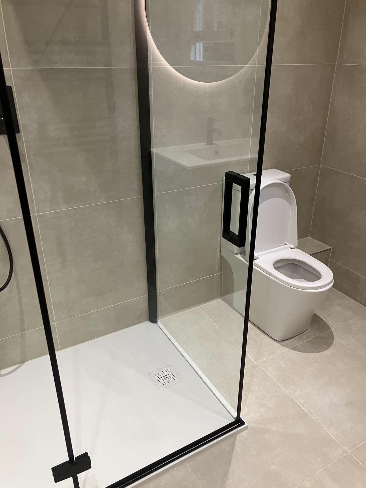 Glass-screened shower with black recessed niche shelving and round backlit mirror reflected in the background