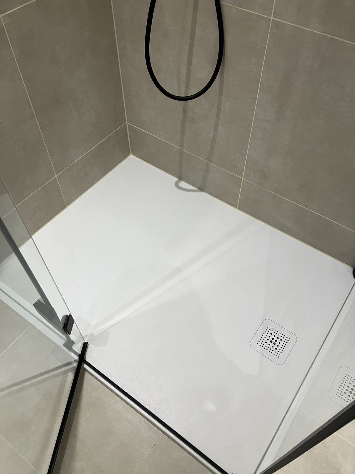 Corner of a walk-in shower tray with black shower hose against stone-effect tiling and a second drain visible