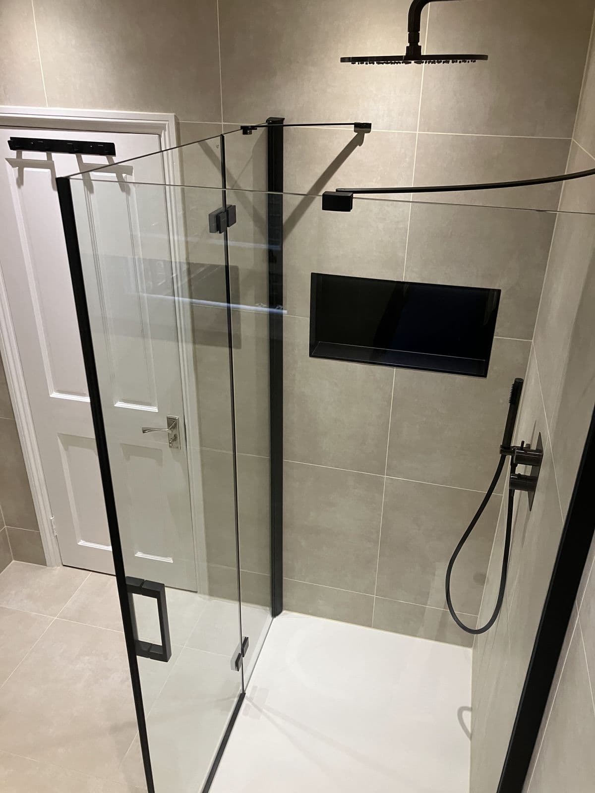 Frameless glass shower door with black hinges opening onto a family bathroom, white panelled door reflected inside