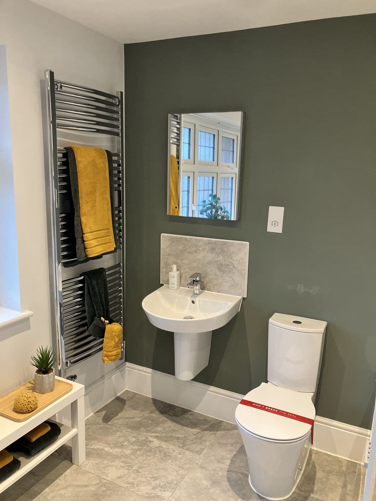 Sage-green bathroom with pedestal basin, square mirror and toilet fitted with protective seal tape, chrome ladder towel rail with mustard and charcoal towels