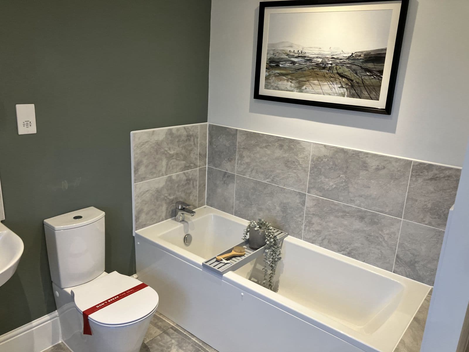 Sage-green bathroom with bath, framed landscape artwork, stone-effect tile splashback and toilet fitted with protective seal tape