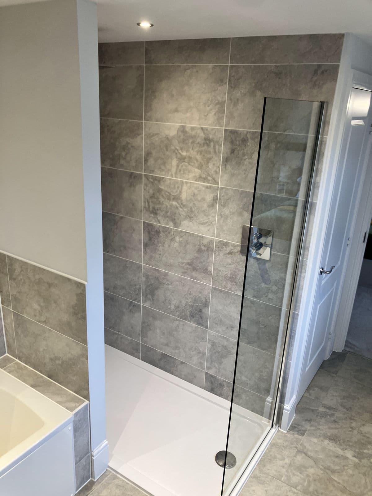 Frameless glass shower screen in a stone-effect tiled corner shower, chrome thermostatic valve, bedroom door visible beyond