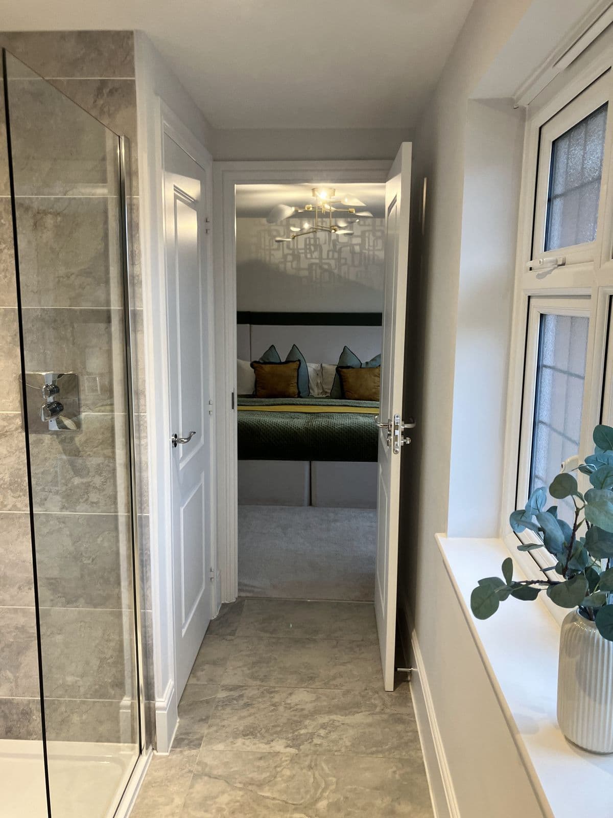 Doorway view from an en-suite shower through to a bedroom with green upholstered headboard and patterned wallpaper
