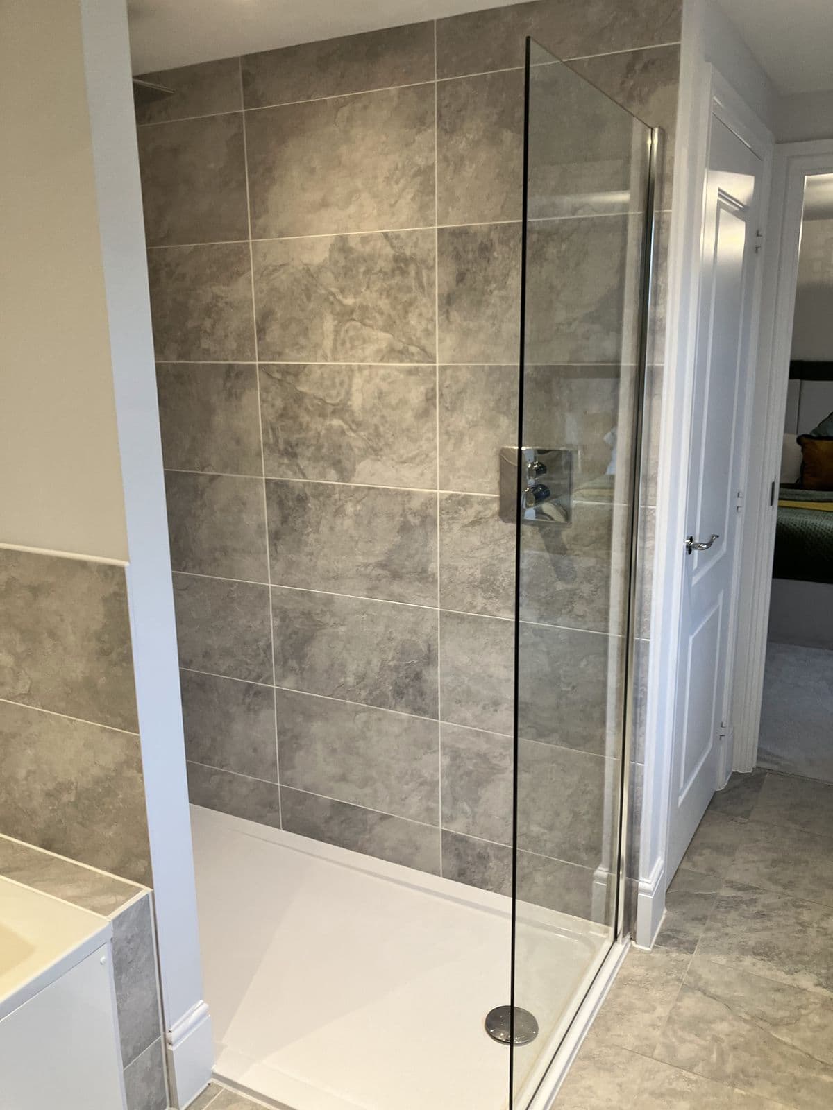 Bath with stone-effect tile splashback beside a walk-in shower with matching tiling and chrome thermostatic valve