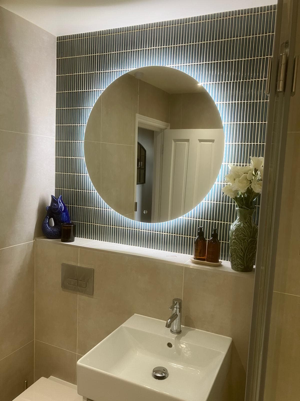 Round backlit mirror above a teal linear-tiled splashback, with basin, chrome mixer tap and decorative ceramic fish ornament