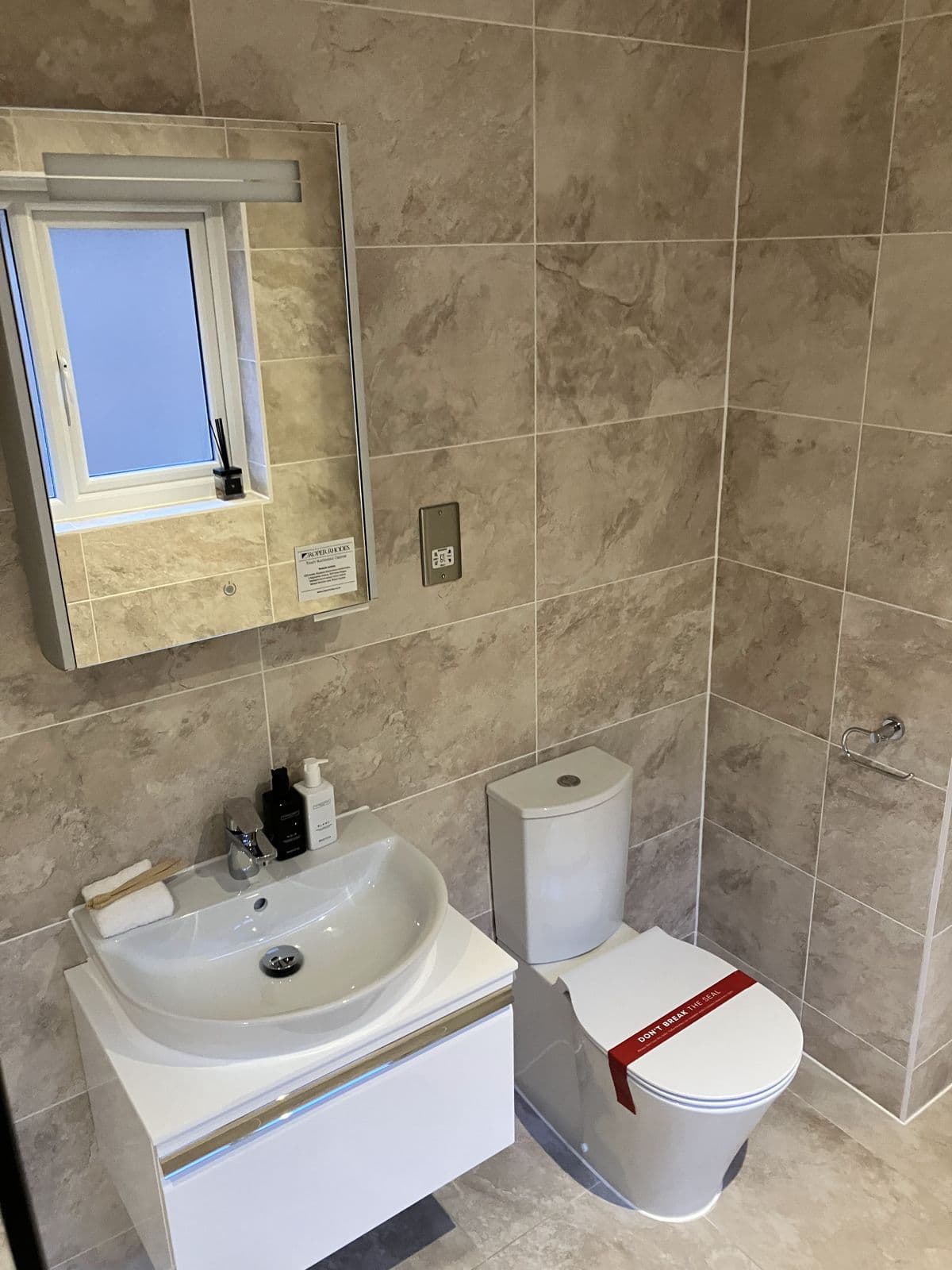 Beige stone-effect tiled bathroom with wall-hung basin, mirrored cabinet and toilet fitted with protective seal tape