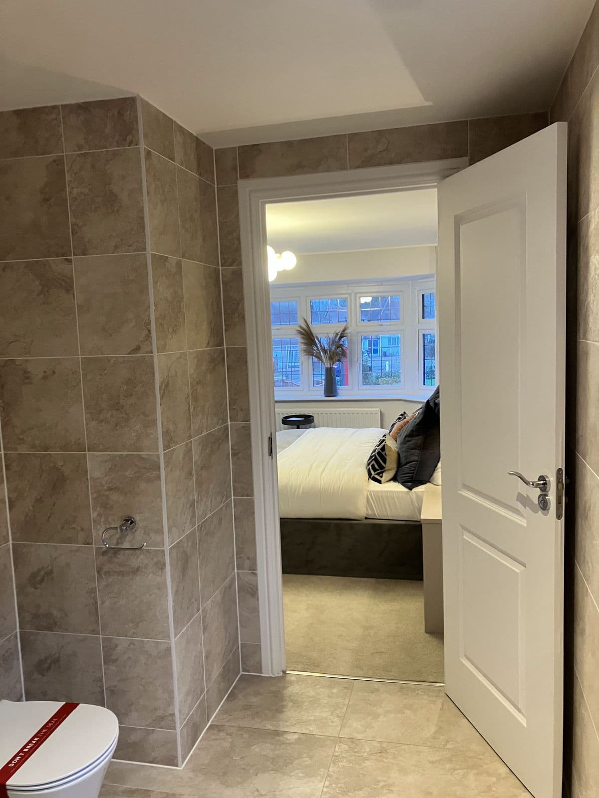 Doorway view from an en-suite through to a bedroom with bay window, pendant lighting and a dried pampas grass arrangement