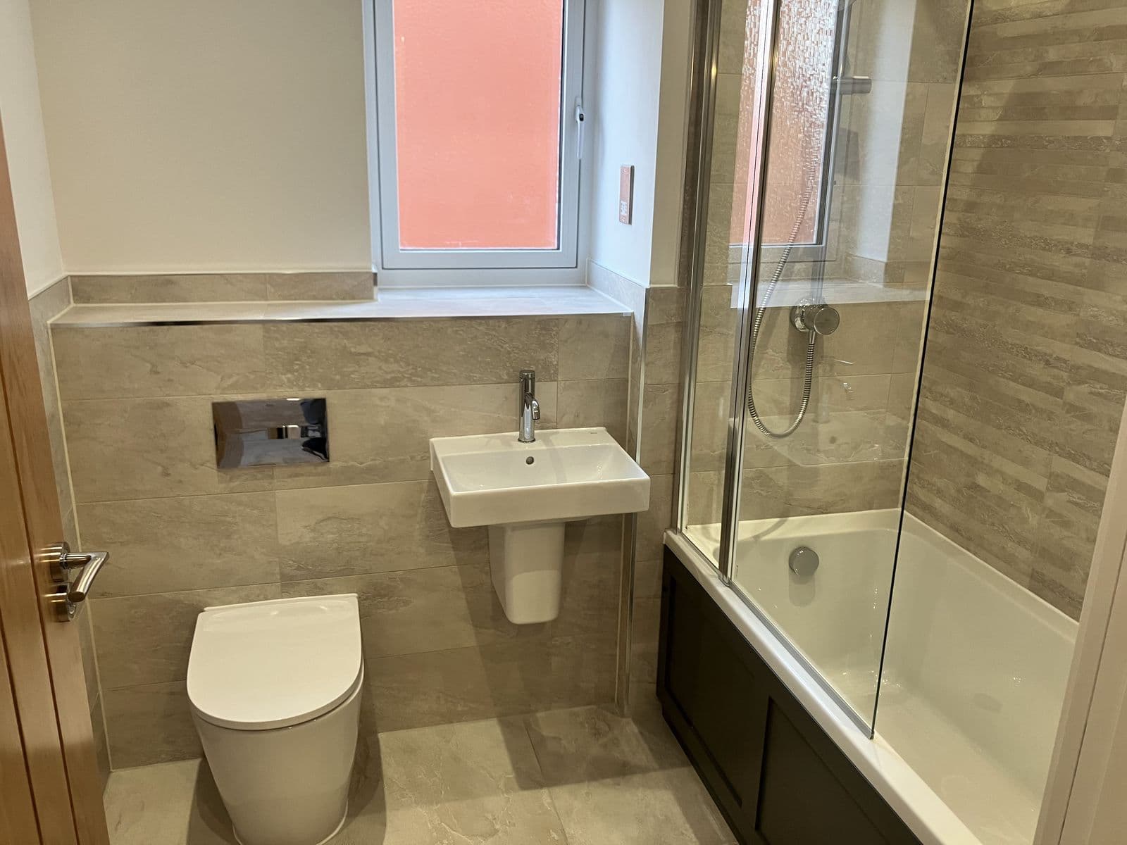 Wall-hung basin and pedestal beneath a coral-tinted obscured window, beside a bath with folding glass shower screen and stone-effect tiling