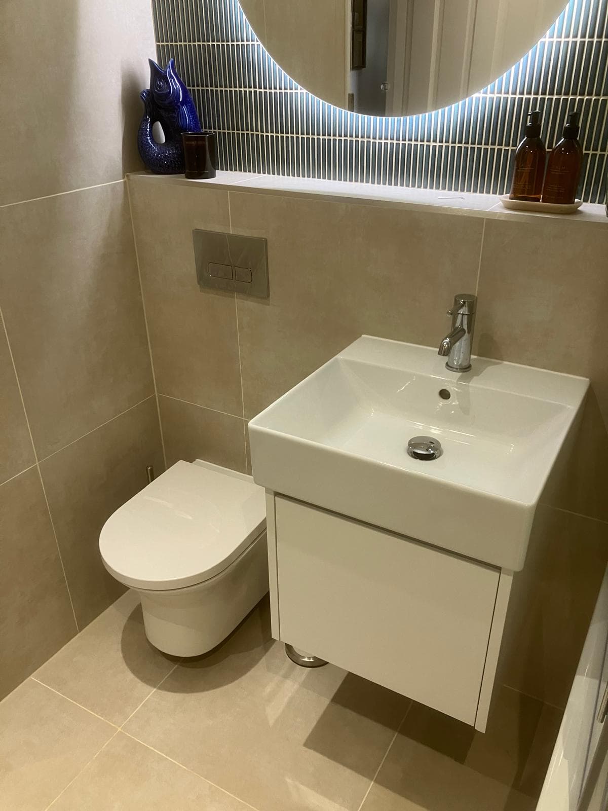 Wall-hung basin and back-to-wall toilet with concealed cistern flush plate beneath a round backlit mirror and blue linear tile