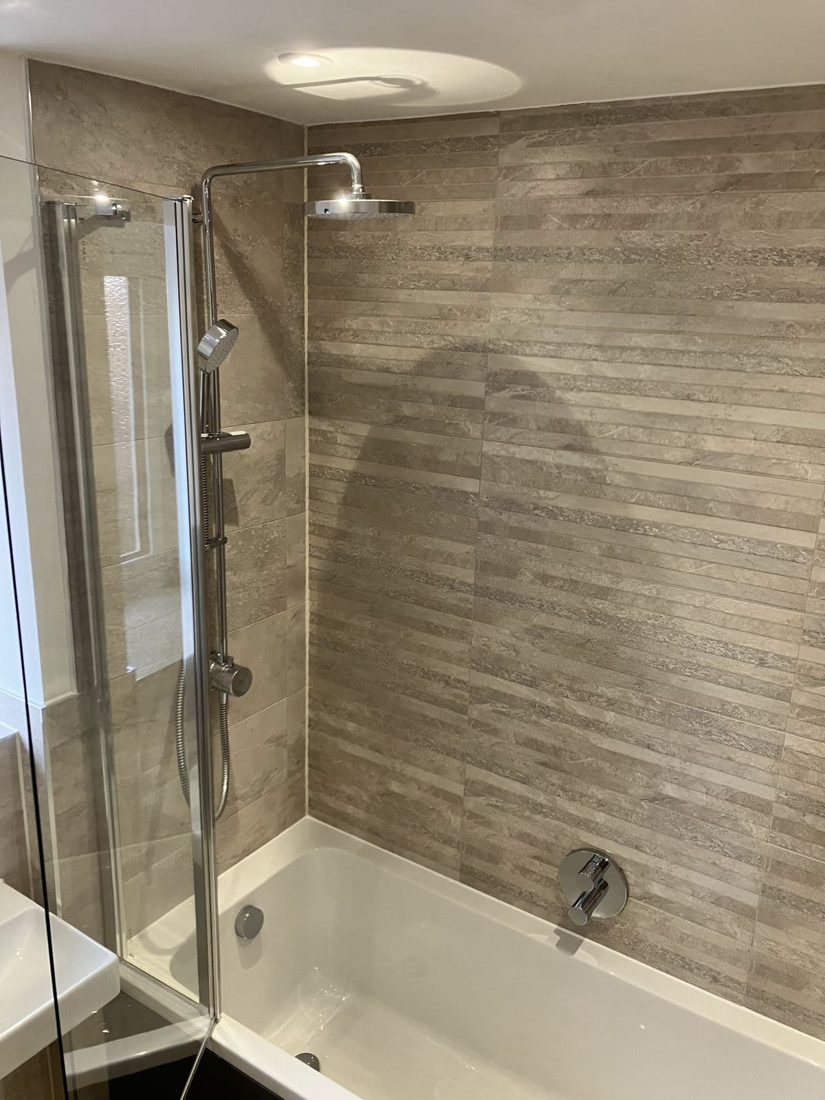 Rainfall shower head and riser rail over a bath, with linear stone-effect tiled feature wall and chrome bath filler