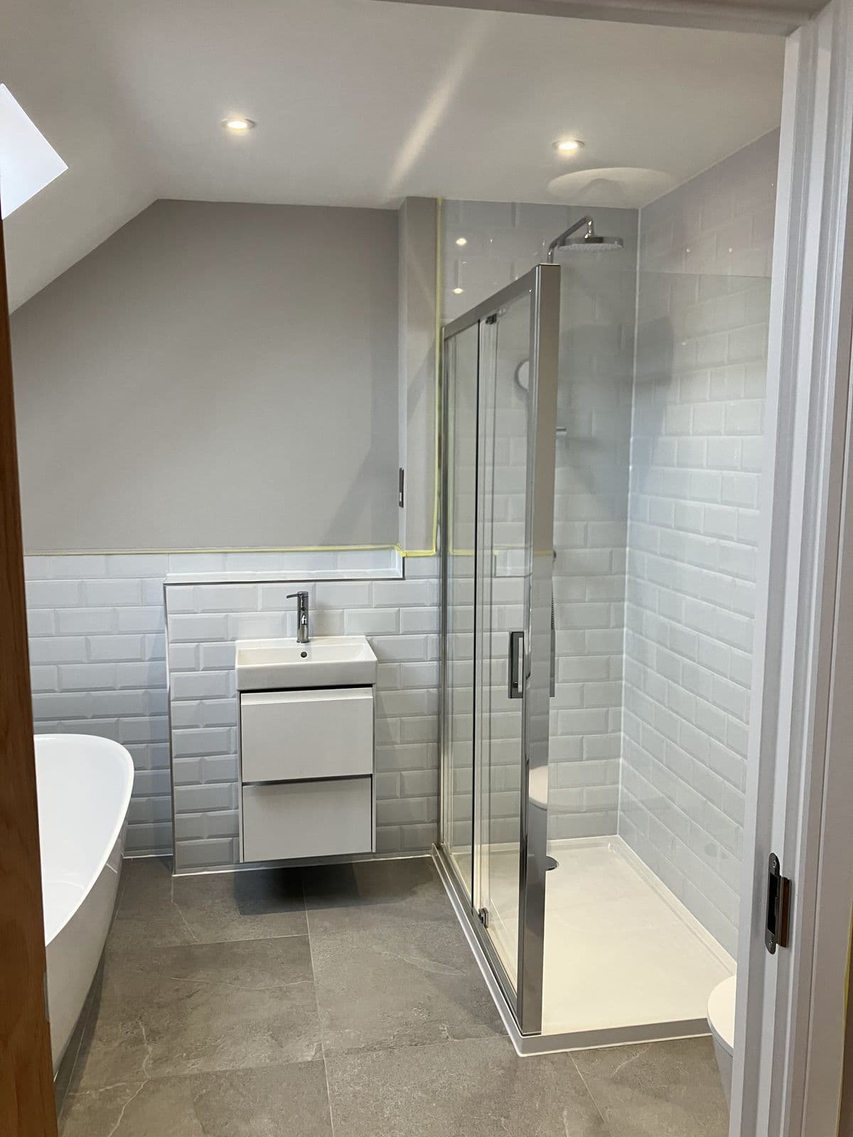 Loft en-suite with grey metro-tiled shower, freestanding bath and wall-hung vanity basin, set under a sloped ceiling