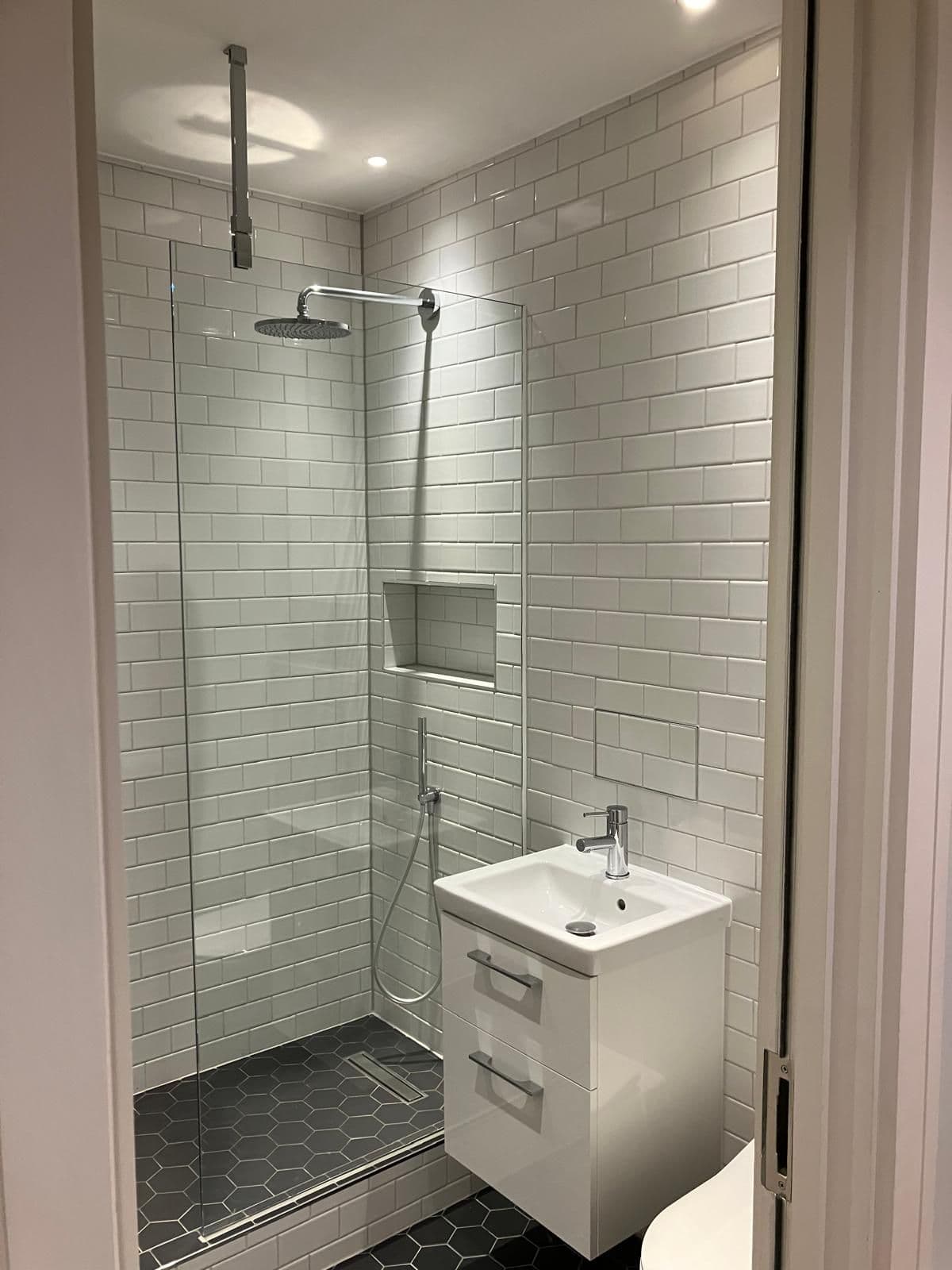 Walk-in shower with hexagonal black floor tiles, white metro wall tiles and a wall-hung basin beside it