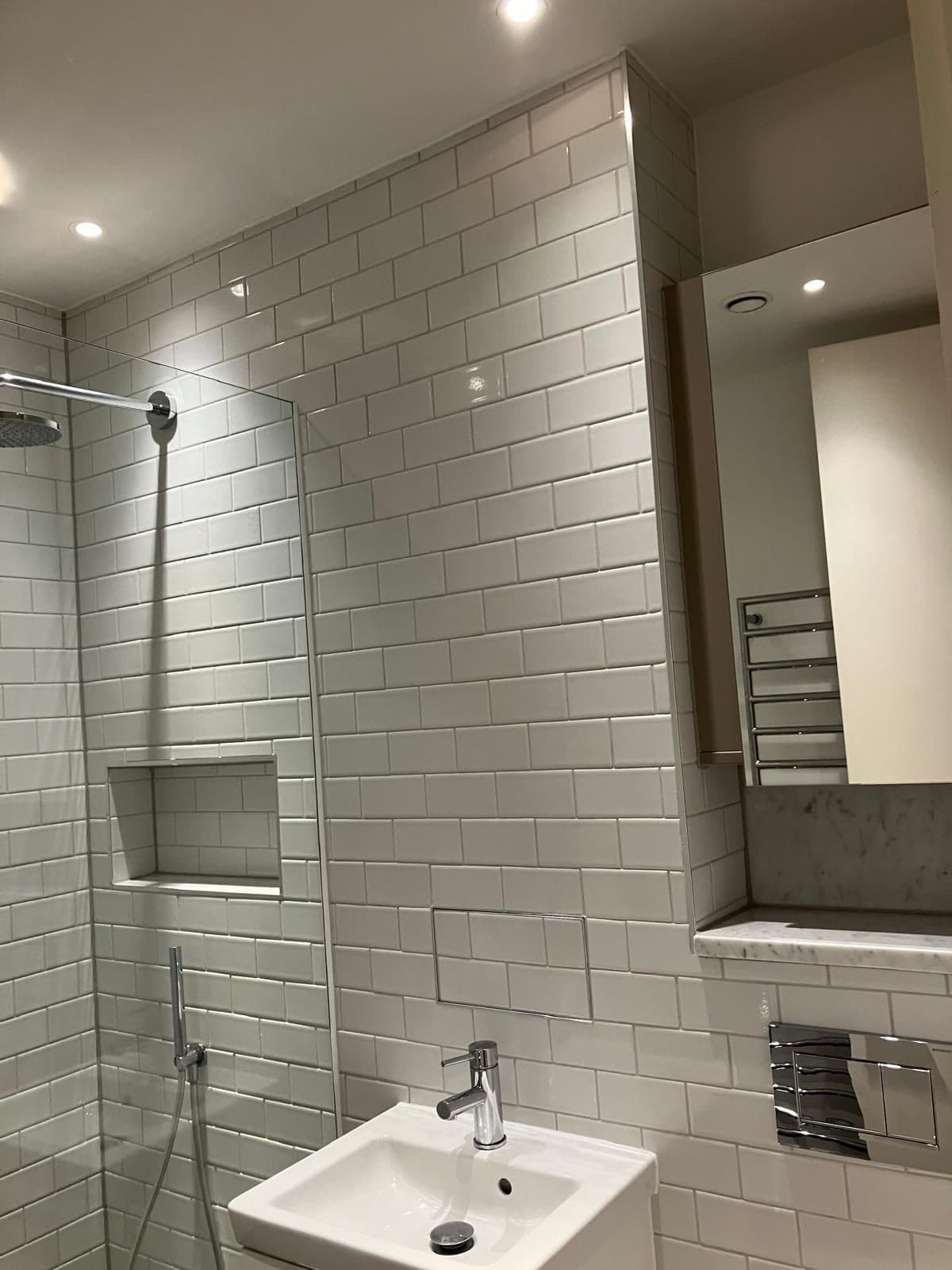 White metro-tiled shower and vanity area with marble-effect display shelf and chrome heated towel rail reflected in the mirror