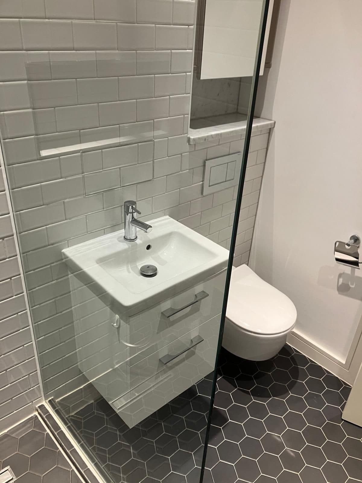 Wall-hung vanity basin and toilet with white metro tiling, black hexagonal floor tiles and marble-effect display shelf