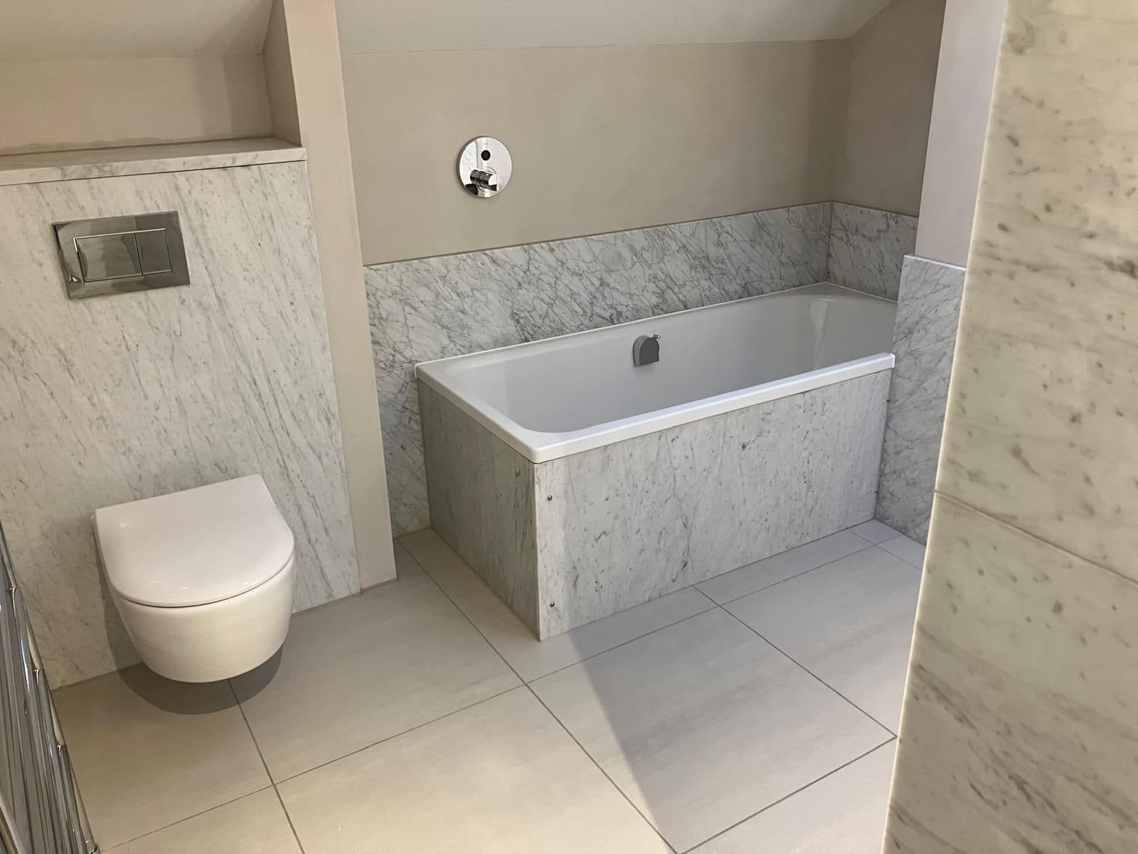 Marble-clad bath panel with wall-mounted chrome bath filler, beside a wall-hung toilet with matching marble tiling