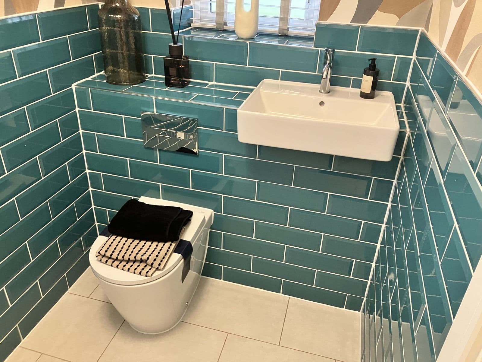 Teal metro-tiled cloakroom with wall-hung basin, chrome mixer tap, reed diffuser and glass bottle vase on a tiled display shelf