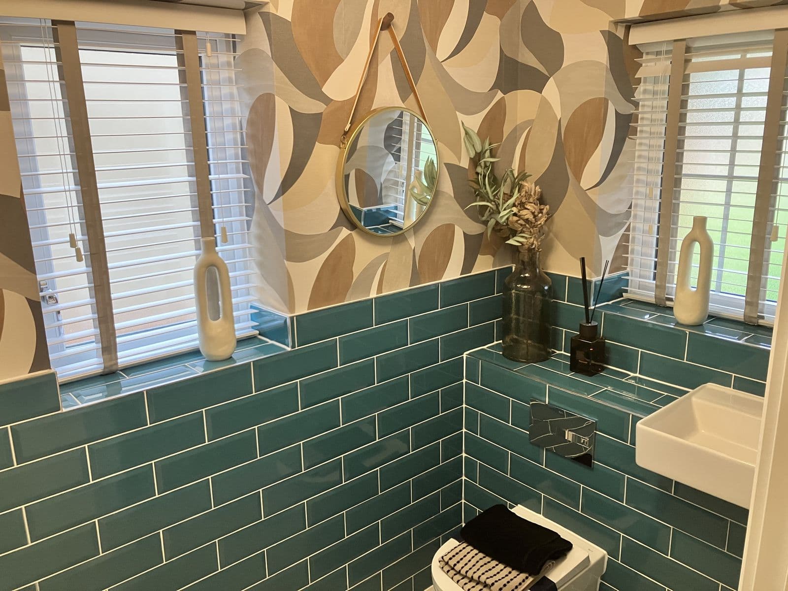 Teal metro-tiled cloakroom window sill styled with a ceramic vase and patterned feature wallpaper, round mirror and basin visible alongside
