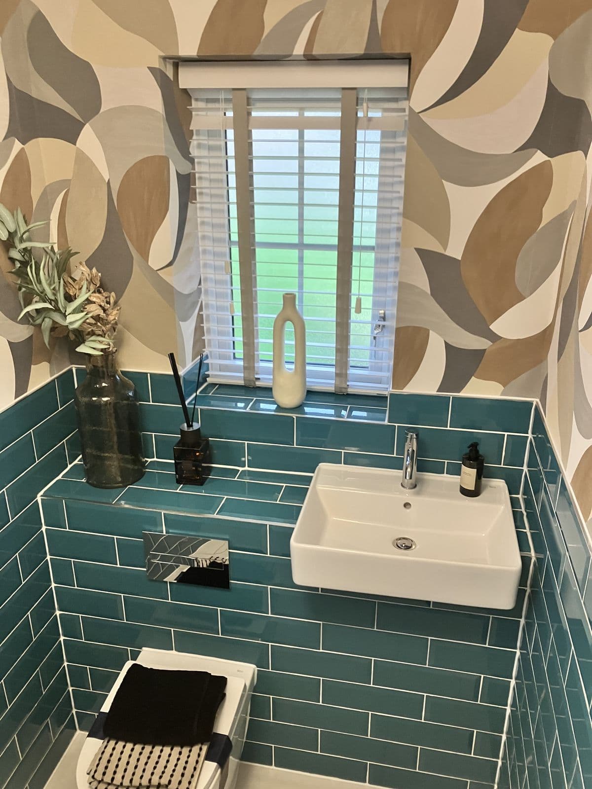 Teal metro-tiled cloakroom, wider view showing the basin, patterned wallpaper and toilet styled with folded towels
