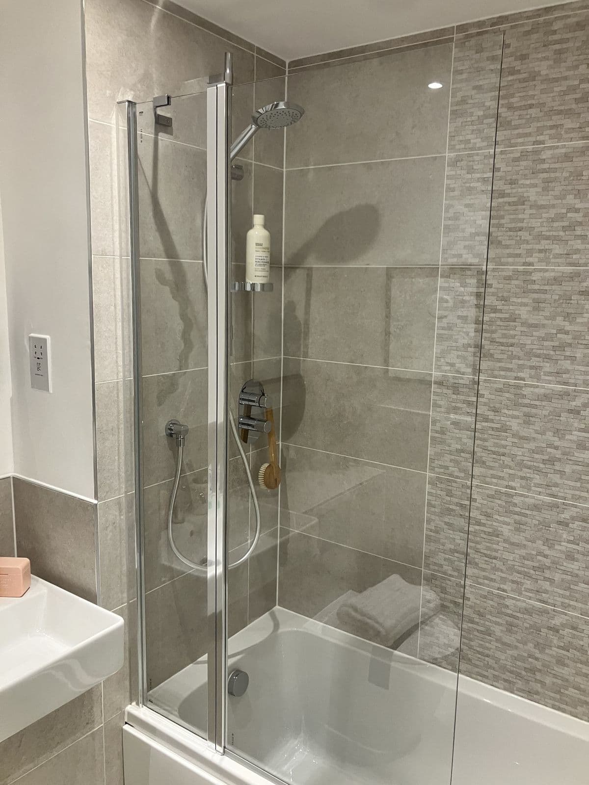 Fold-down glass shower screen over a bath, with a wall-mounted shower gel holder, thermostatic valve and shower brush