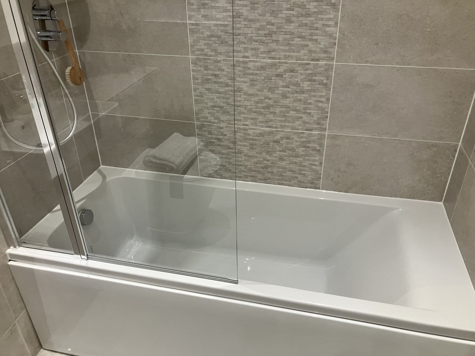 Bath with a folded towel and mixed stone-effect mosaic feature tile, seen through the folding glass shower screen