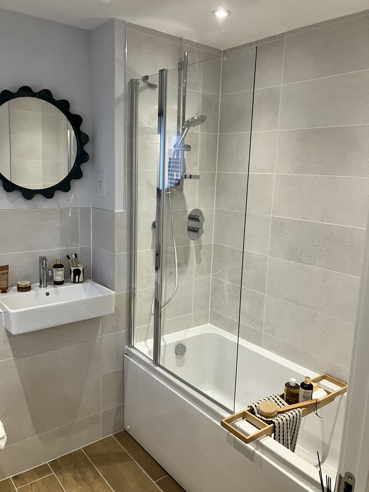 Fold-down glass bath screen with riser-rail shower, wall-hung basin and a wooden bath caddy with brush and bottles