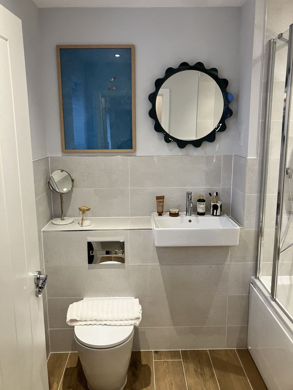 Wall-hung basin styled with skincare products beneath a scalloped black mirror, small round vanity mirror and framed blue artwork alongside