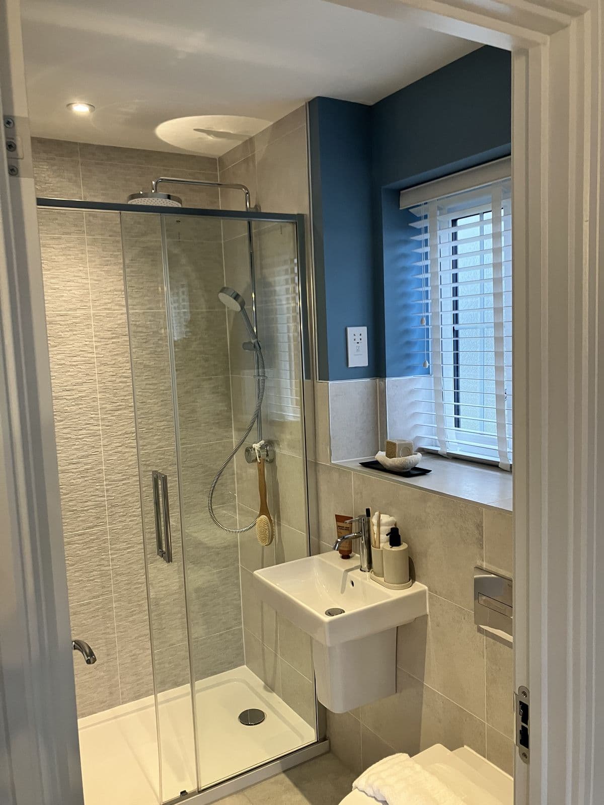 Doorway view into a sliding-door shower enclosure and wall-hung basin, with a deep blue accent wall and dressed window beyond