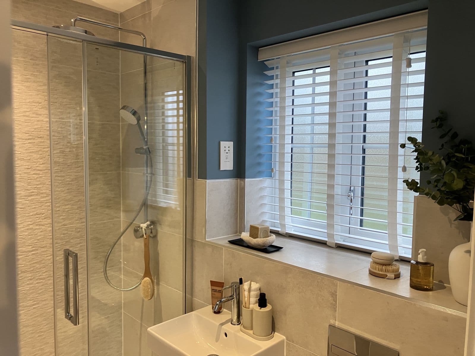 Close-up of a windowsill styled with soap dish, body brush and reed diffuser beside a wall-hung basin with toiletries and concealed cistern flush plate